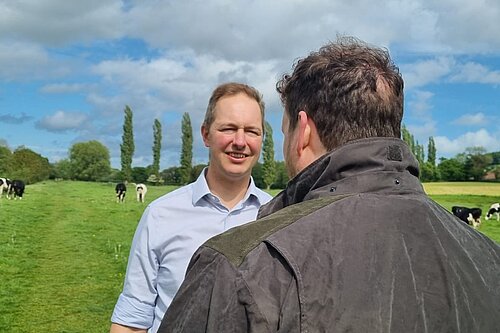 A photo of Richard Foord talking to someone whose back is to the camera, and both people are in a field with cows in the background