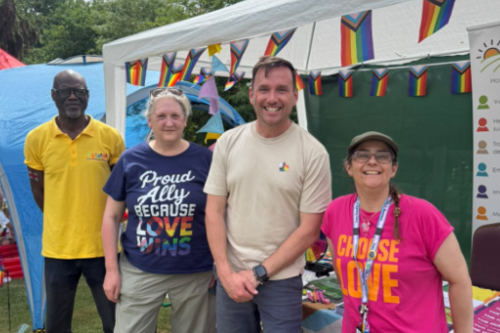 Sean in stood in a group of people (including councillor Liz Needham to his right) in front of a gazebo decorated with Pride flag bunting.  