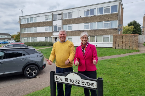 Two people (Tony on the left, Daisy on the right) are standing in front of a block of flats, and behind a sign displaying flat numbers. They are both smiling, looking directly at the camera, and giving a thumbs up.