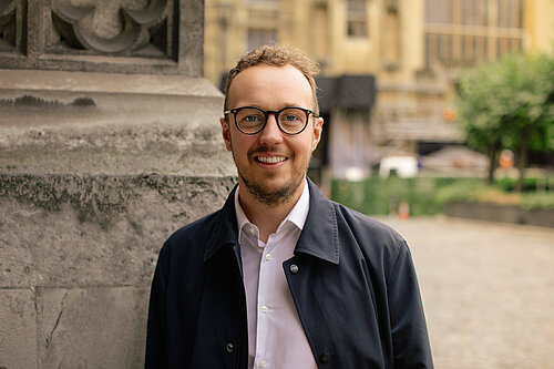 A man wearing glasses stands outdoors beside a stone wall, smiling at the camera.