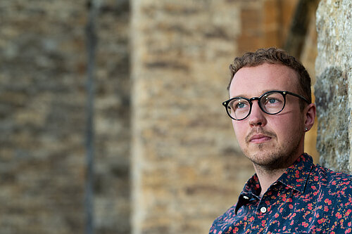 Adam Dance MP stands against a brick wall, looking serious. 